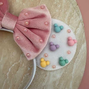 May include: Pink velvet bow headband with pearl accents. The headband features a white circular accent with colorful Mickey Mouse-shaped designs. A fun and whimsical accessory.