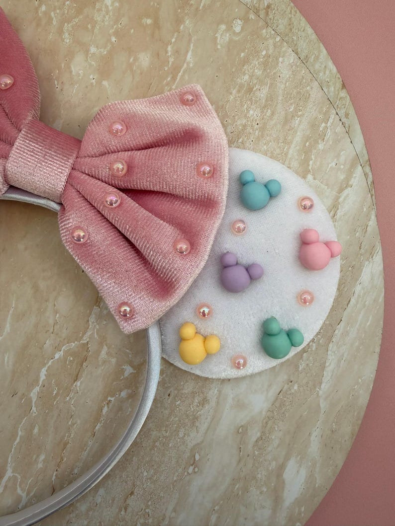 May include: Pink velvet bow headband with pearl accents. The headband features a white circular accent with colorful Mickey Mouse-shaped designs. A fun and whimsical accessory.