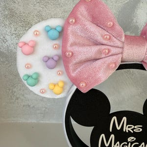 May include: A white headband with black Mickey Mouse ears and a pink bow. The left ear features colorful Mickey Mouse shapes and pink pearls. The bow is adorned with pink pearls. The text "Mrs Magical Ears" is printed on the right ear.
