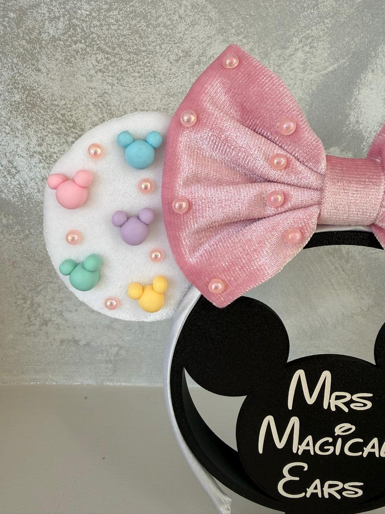 May include: A white headband with black Mickey Mouse ears and a pink bow. The left ear features colorful Mickey Mouse shapes and pink pearls. The bow is adorned with pink pearls. The text "Mrs Magical Ears" is printed on the right ear.
