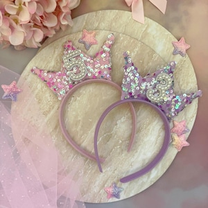 May include: Two sequined crown headbands, one pink and one purple, each with a number 5. The crowns are decorated with sequins and pearls. The headbands are on a marble-effect surface, surrounded by star-shaped embellishments and pink tulle.