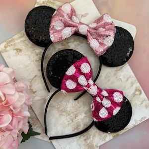 May include: Two sequined mouse ear headbands. One has a pink bow with white polka dots, the other has a fuchsia bow with white polka dots. The ears are black and the headbands are black.