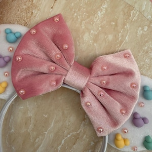 May include: A pink velvet bow headband with pearl accents. The headband features white ears decorated with colorful, cartoon-like figures. The bow is centered on a white plastic band, creating a playful accessory.