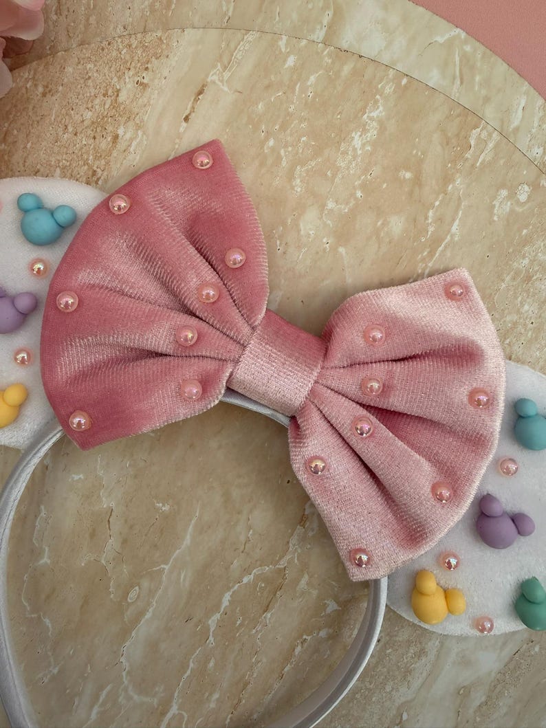May include: A pink velvet bow headband with pearl accents. The headband features white ears decorated with colorful, cartoon-like figures. The bow is centered on a white plastic band, creating a playful accessory.