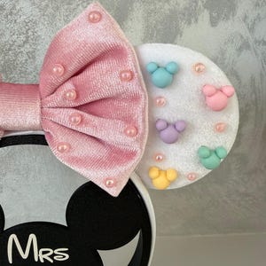 May include: A pink velvet bow headband with pearl accents. The headband features a white ear with colorful mouse-shaped embellishments and the text "Mrs Magical".