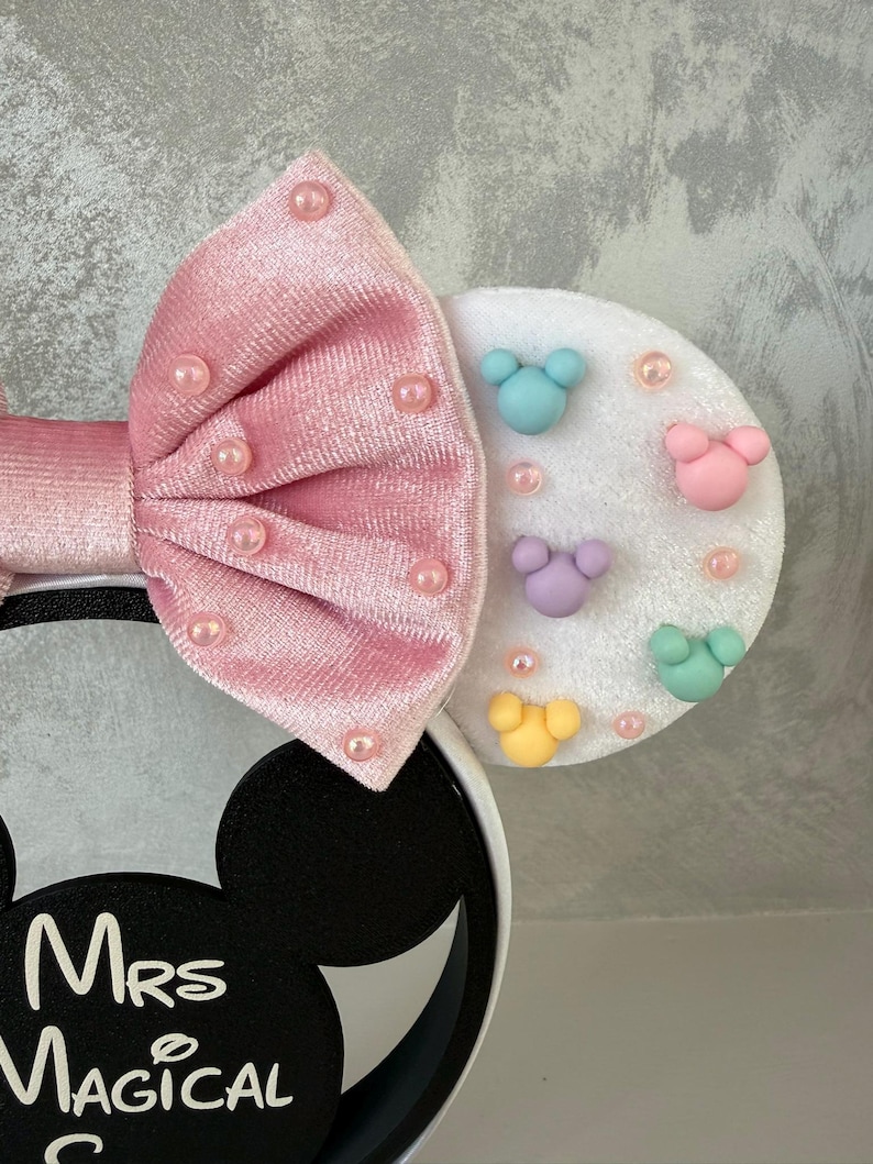 May include: A pink velvet bow headband with pearl accents. The headband features a white ear with colorful mouse-shaped embellishments and the text "Mrs Magical".