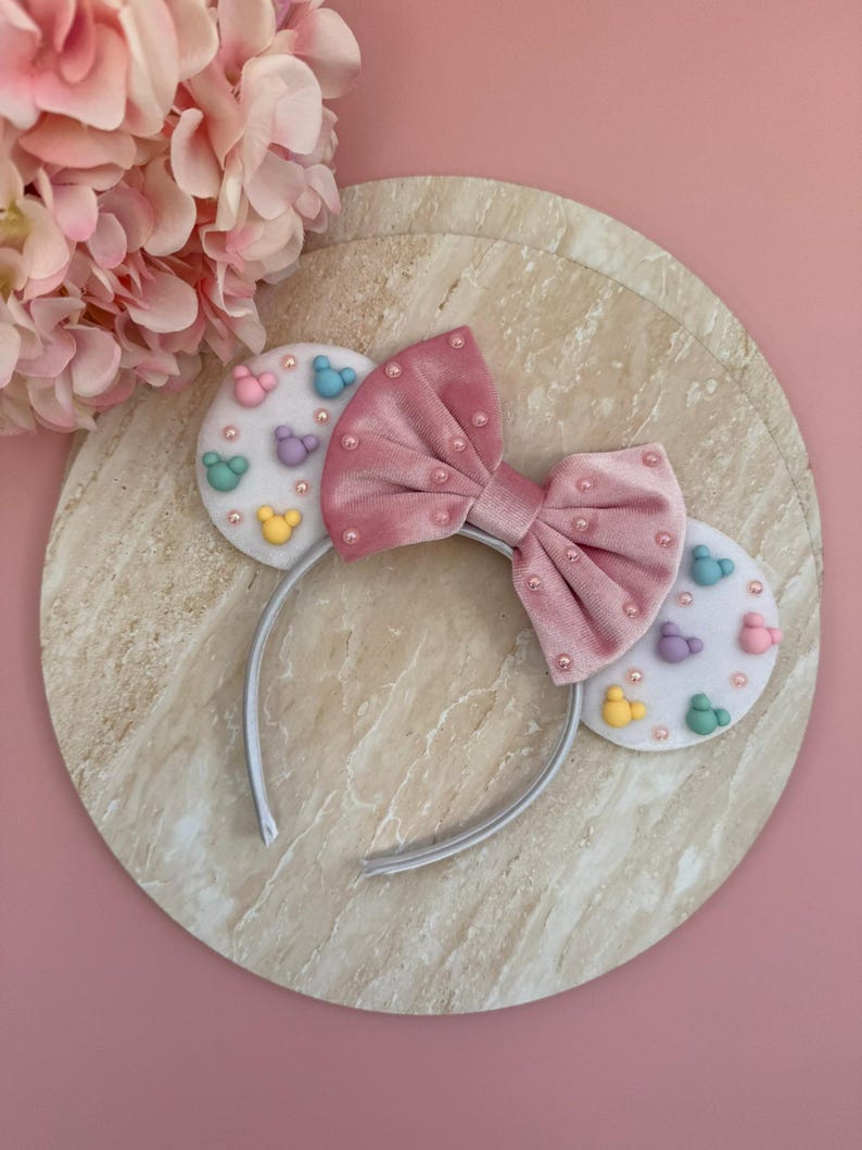 May include: A pink velvet bow headband with white ears decorated with colourful mouse silhouettes. The bow is adorned with small pink beads. The headband is on a marble-like surface against a pink background.