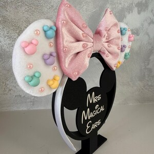 May include: A black Mickey Mouse ear headband with a pink velvet bow and white ears. The ears are decorated with pastel-colored Mickey Mouse shapes and pearls. The base reads "Mrs Magical Ears".