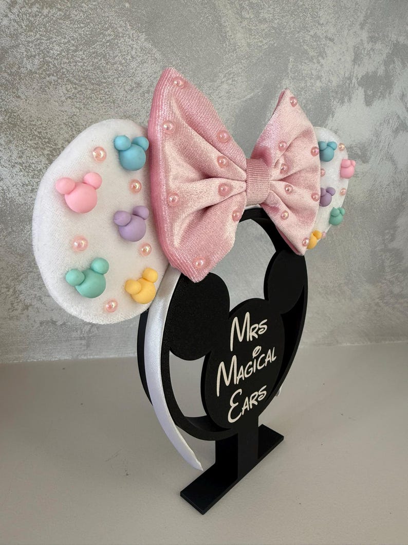 May include: A black Mickey Mouse ear headband with a pink velvet bow and white ears. The ears are decorated with pastel-colored Mickey Mouse shapes and pearls. The base reads "Mrs Magical Ears".