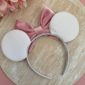 May include: A white headband with two large, round, white plush ears. A pink velvet bow is centered between the ears. The headband is white and the bow is a dusty rose color. The background is a marble-like surface.