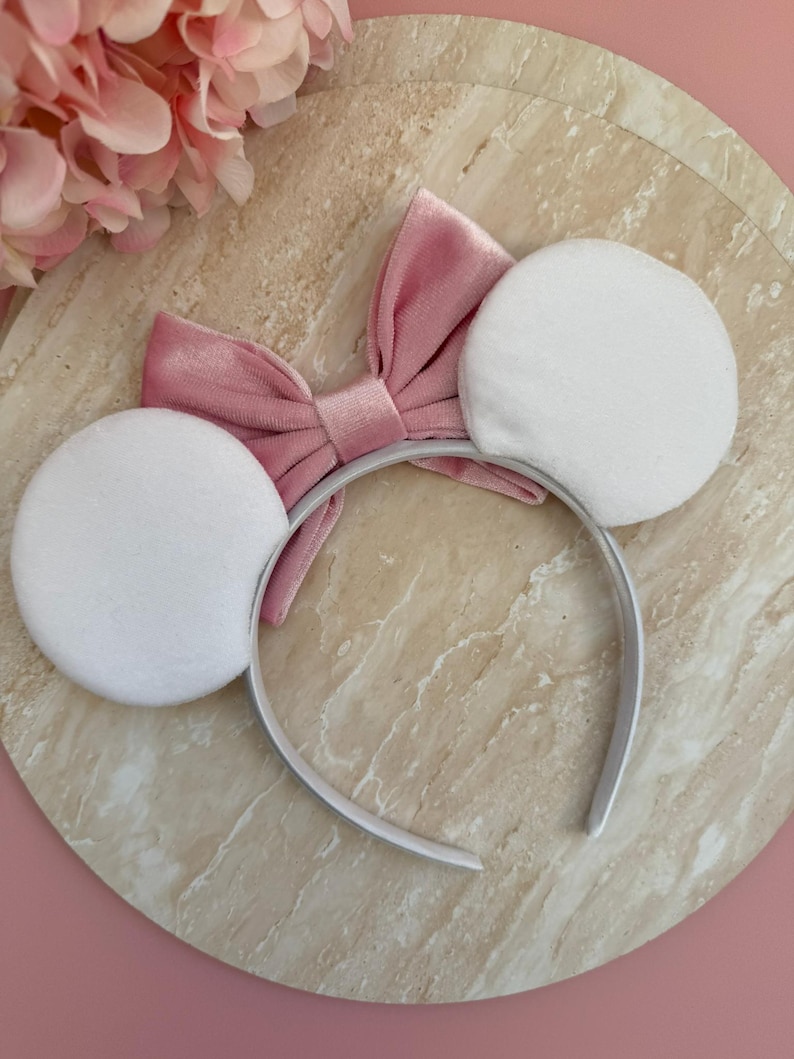 May include: A white headband with two large, round, white plush ears. A pink velvet bow is centered between the ears. The headband is white and the bow is a dusty rose color. The background is a marble-like surface.