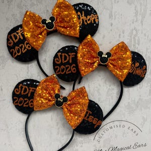 May include: Black mouse ear headbands with orange sequin bows and black sequin ears. The ears have custom text, including initials and the year 2026. The bows have a black Mickey Mouse emblem in the centre.