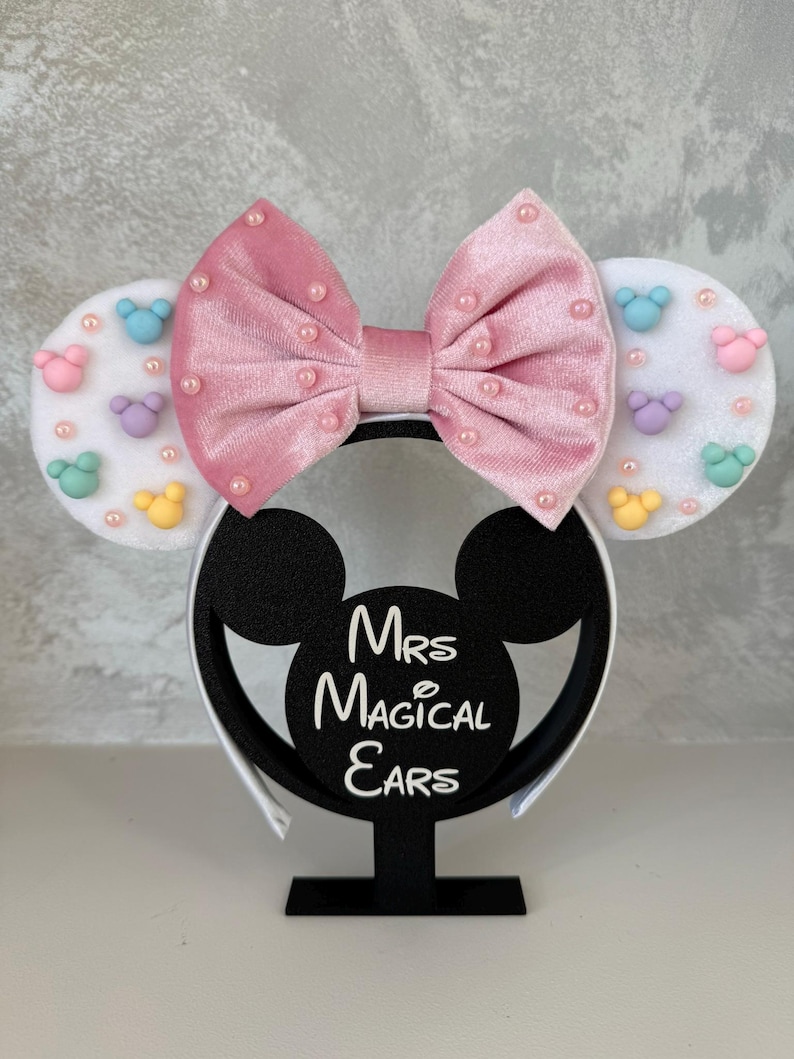 May include: A pink velvet bow headband with pearl accents and white ears decorated with pastel-colored mouse shapes. The headband is displayed on a black stand with the text "Mrs Magical Ears".