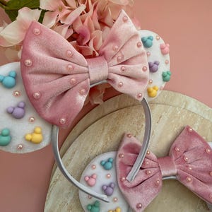 May include: Pink velvet bow headband with pearl accents. The headband features white circular accents with colorful cartoon character designs. The background includes pink flowers and a light-colored surface.