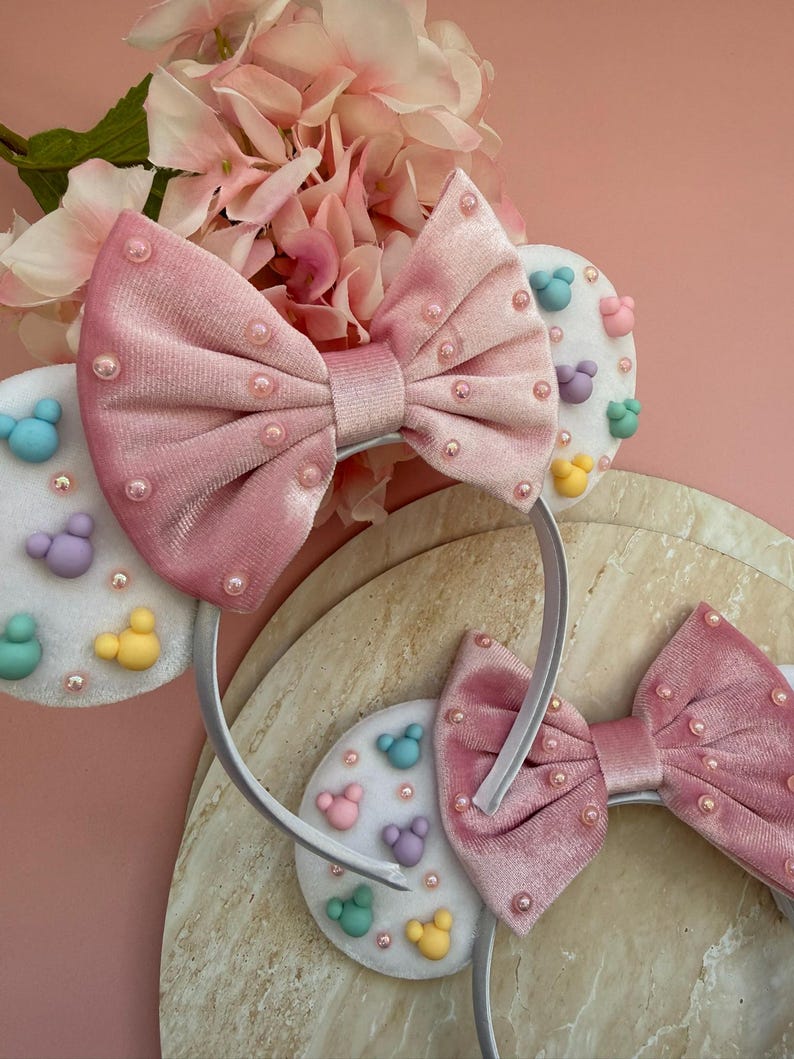 May include: Pink velvet bow headband with pearl accents. The headband features white circular accents with colorful cartoon character designs. The background includes pink flowers and a light-colored surface.