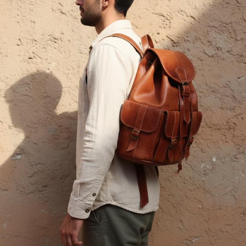 Man wearing handmade brown leather backpack, vintage travel rucksack with buckle pockets and adjustable straps, rustic leather bag for work, hiking, college and everyday carry