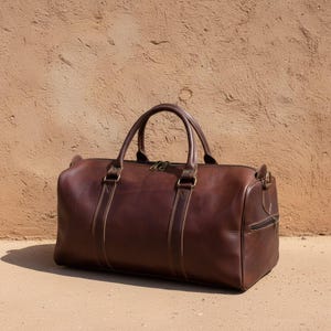 Puede incluir: Una bolsa de lona de cuero marrón con dos asas superiores y un bolsillo lateral con cremallera. La bolsa tiene forma rectangular y está hecha de cuero liso y pulido. La bolsa está sobre una superficie de hormigón.