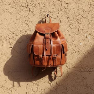 Handmade brown leather backpack hanging on textured wall, vintage travel rucksack with buckle pockets and rustic boho style for everyday carry and adventure