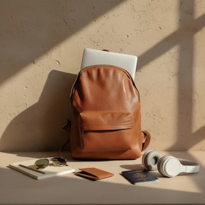 Pode incluir: Uma mochila de couro castanho com um laptop dentro, encostada a uma parede bege. Ao lado, óculos de sol, um caderno com um lápis, uma carteira pequena, um passaporte e fones de ouvido brancos.