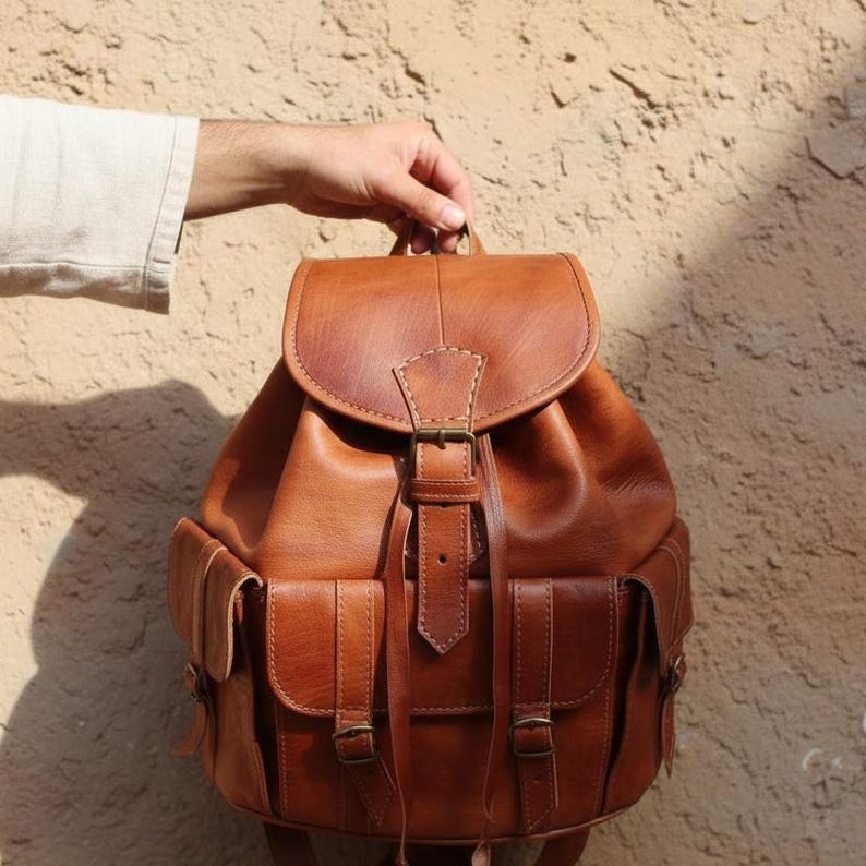 Handmade vintage brown leather backpack held by hand, rustic travel rucksack with buckle straps and front pockets, boho leather bag for work, college, hiking and everyday carry