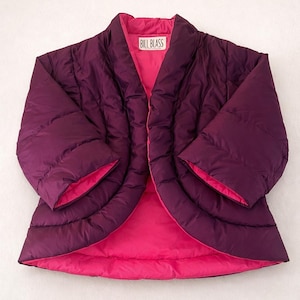 May include: A reversible quilted jacket in a deep purple with a bright pink lining. The jacket has a shawl collar and wide sleeves. The label "BILL BLASS" is visible on the inside collar. The jacket is designed with a relaxed fit.
