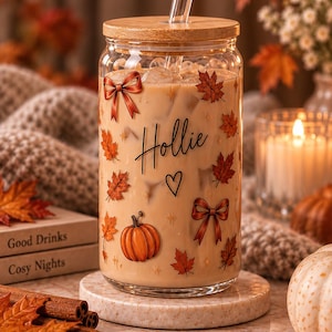 Puede incluir: Un vaso transparente con tapa de bambú y pajita de cristal, lleno de una bebida helada de color beige. El exterior presenta un diseño impreso de calabazas naranjas, hojas de arce rojas, lazos y el nombre Hollie en letra negra.