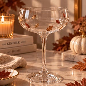 Op de afbeelding: Een helder glazen coupe cocktailglas met een delicaat patroon van kleine, metaalkleurige herfstbladeren verspreid over de kom. Het glas heeft een slanke steel en een brede, ronde voet met een subtiele textuur van concentrische ringen.