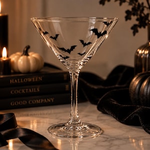 Puede incluir: Una copa de martini de cristal transparente con un patrón de pequeñas siluetas de murciélagos negros impresas alrededor del exterior del cuenco. El tallo es delgado y termina en una base circular, proporcionando estabilidad a la copa.