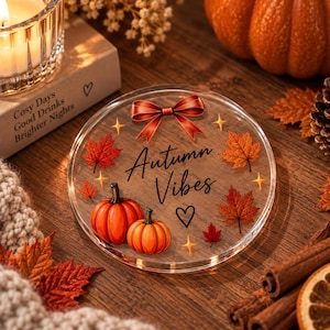 Könnte beinhalten: Ein runder, klarer Acryl-Untersetzer mit einer roten Schleife oben, zwei orangefarbenen Kürbissen unten sowie verstreuten Ahornblättern und goldenen Sternen. In der Mitte steht der Text Autumn Vibes in schwarzer Schrift mit einem Herz.