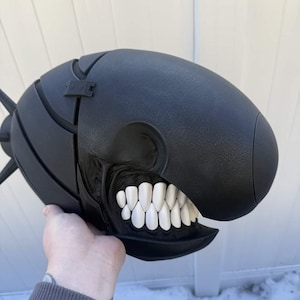Könnte beinhalten: Eine schwarze, stilisierte Maske mit einem großen, offenen Mund, der weiße Zähne zeigt. Die Maske hat eine abgerundete, ovale Form mit einer strukturierten Oberfläche und einem kleinen, rechteckigen Aufsatz oben. Die Maske wird von einer Person gehalten.