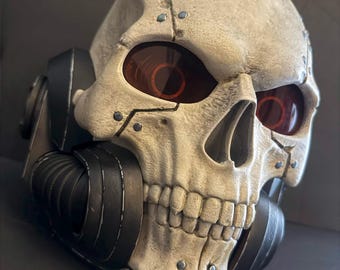 Könnte beinhalten: Eine detaillierte Totenkopfmaske mit roten Augenlinsen und schwarzen Akzenten. Die Maske hat ein verwittertes, knochenartiges Aussehen mit sichtbaren Nieten und einem Atemschutzmasken-Design. Das Gesamtbild deutet auf ein postapokalyptisches oder Science-Fiction-Thema hin.