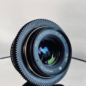 Puede incluir: Un objetivo de cámara negro con el texto "MC Гелиос-77M-4" y "1.8/50" impreso. El objetivo tiene un anillo exterior texturizado y estriado. El objetivo se refleja en una superficie brillante.