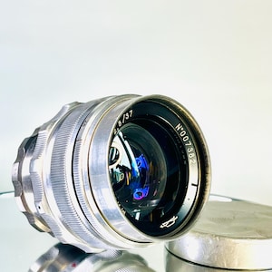May include: A vintage silver camera lens with the markings "2.8/37" and "N°007382". The lens has a ribbed design and a reflective surface, with a lens cap visible in the background. The lens is likely for a classic or antique camera.