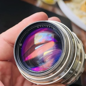 Può includere: Un obiettivo per fotocamera argentato e nero con il testo "ЮПИТЕР-9" visibile. L'obiettivo ha una superficie riflettente con rifrazioni di luce colorate. L'obiettivo è tenuto in mano.