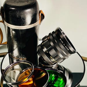 May include: A black camera lens with "Made in USSR" text, resting on a reflective surface, alongside a black leather case and several colored lens filters. The filters include clear, orange, and green.