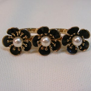 May include: A gold-toned ring featuring three black flower designs with pearl centers. The flowers have detailed petals and are set on a thin band. The ring is displayed on a white surface.