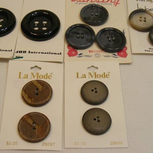May include: Assortment of buttons in various colors and sizes, including black, gray, and brown. Some buttons are attached to cards with brand names like "La Mode" and "JHB International".