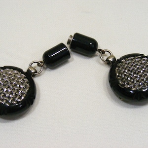 May include: A pair of vintage black and silver tone earrings. Each earring features a round, black, carved frame with a silver-colored chainmail design in the center. The earrings are connected by a silver-colored chain and a black cylindrical clasp.