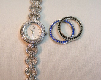 May include: A silver watch with a white face and a crystal-encrusted band. The watch is accompanied by two silver rings with blue and black crystals.