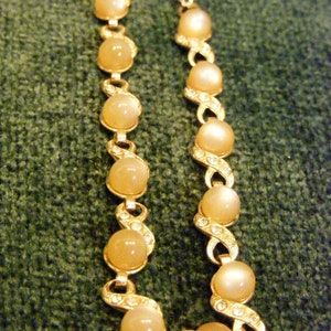 May include: A gold chain necklace with a series of oval-shaped, light brown stones. The stones are set in a gold frame with small, clear rhinestones.