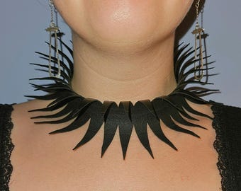 May include: A black leather choker necklace with a spiky, layered design. The necklace is worn with silver earrings. The person is wearing a black top with lace detailing.