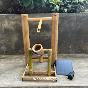 Könnte beinhalten: Bambus-Wasserbrunnen mit Solarpanel und Pumpe. Der Brunnen hat einen natürlichen Bambusrahmen mit Ausguss und einer kleinen Bambusschale. Das Solarpanel und die Pumpe sind schwarz und blau.