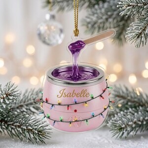 May include: A pink wax warmer ornament featuring a gold string for hanging. The warmer has a metallic rim, a dial, and a wooden applicator stick lifting purple wax. The name Isabelle is printed in gold script, and a string of colorful lights wraps around.