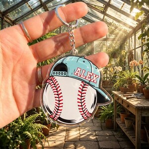 May include: A baseball-themed keychain featuring a baseball with red stitching and a light blue baseball cap. The cap has the name "ALEX" in red letters. The keychain is held up in front of a greenhouse.