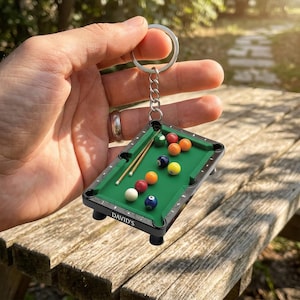 May include: Miniature pool table keychain with a green felt surface, black frame, and colorful billiard balls. Includes two small cues and the text "DAVID'S" on the side. The keychain is held by a silver chain and ring.
