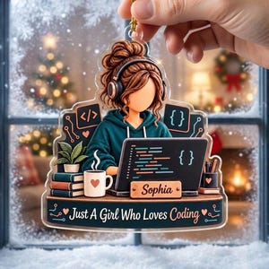 May include: A clear acrylic ornament featuring an illustration of a person with brown hair wearing headphones, working on a laptop with code on the screen. It includes a mug, books, a plant, and the text "Sophia" and "Just A Girl Who Loves Coding."