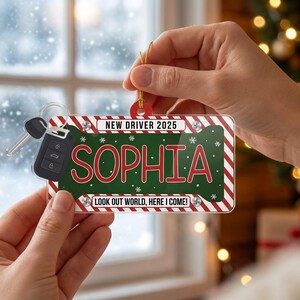 May include: A rectangular acrylic ornament shaped like a license plate with red and white striped borders. The center is green with white snowflakes and the name SOPHIA in red. Text reads NEW DRIVER 2025 and LOOK OUT WORLD, HERE I COME! with a car key.