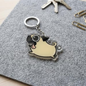 May include: A novelty keychain featuring a cartoon pug dog with a light tan body, black ears and muzzle, and a pink tongue sticking out. The pug is depicted in motion, with a small cloud graphic indicating a fart. The keychain has a silver ring.