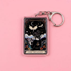 May include: A clear acrylic keychain featuring a tarot card design with a silver-colored metal clasp. The card depicts a woman reading a book under a night sky with a crescent moon and bats. The text "THE READER" is at the bottom.