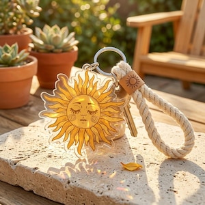 May include: A clear acrylic keychain featuring a yellow sun design with closed eyes and a smiling face. The keychain has a beige rope handle and a small leather tag with a sun emblem. Several keys are attached to the keyring.